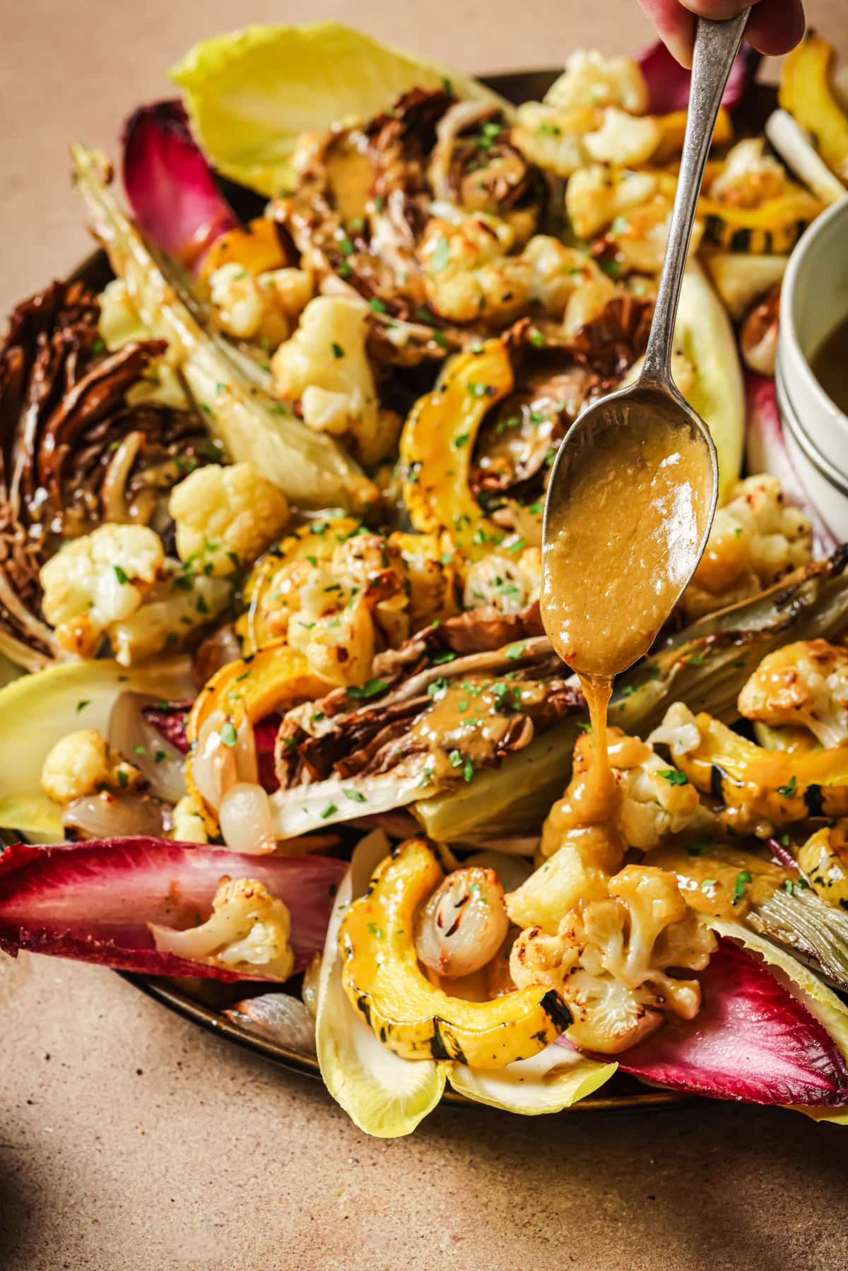 A spoon drizzles dressing over a roasted vegetable salad of cauliflower, squash, and radicchio.