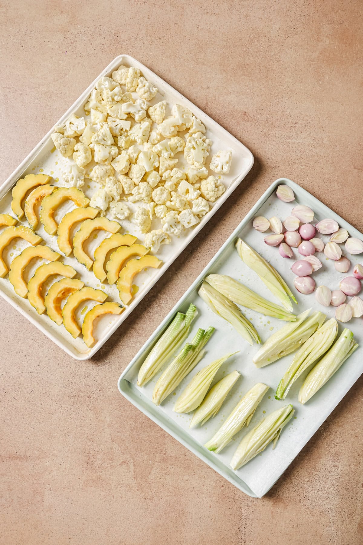 Two baking sheets filled with fresh produce: one holding cauliflower florets and sliced acorn squash, the other holding halved fennel bulbs and whole shallots.