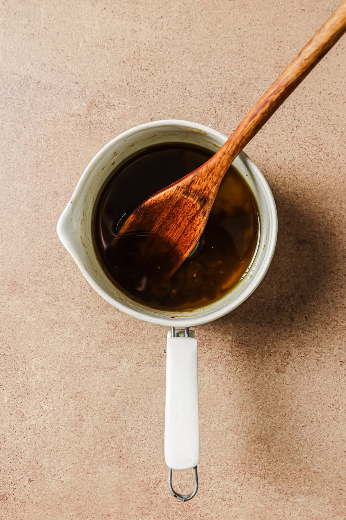A wooden spoon stirs a dark sauce in a small white saucepan.