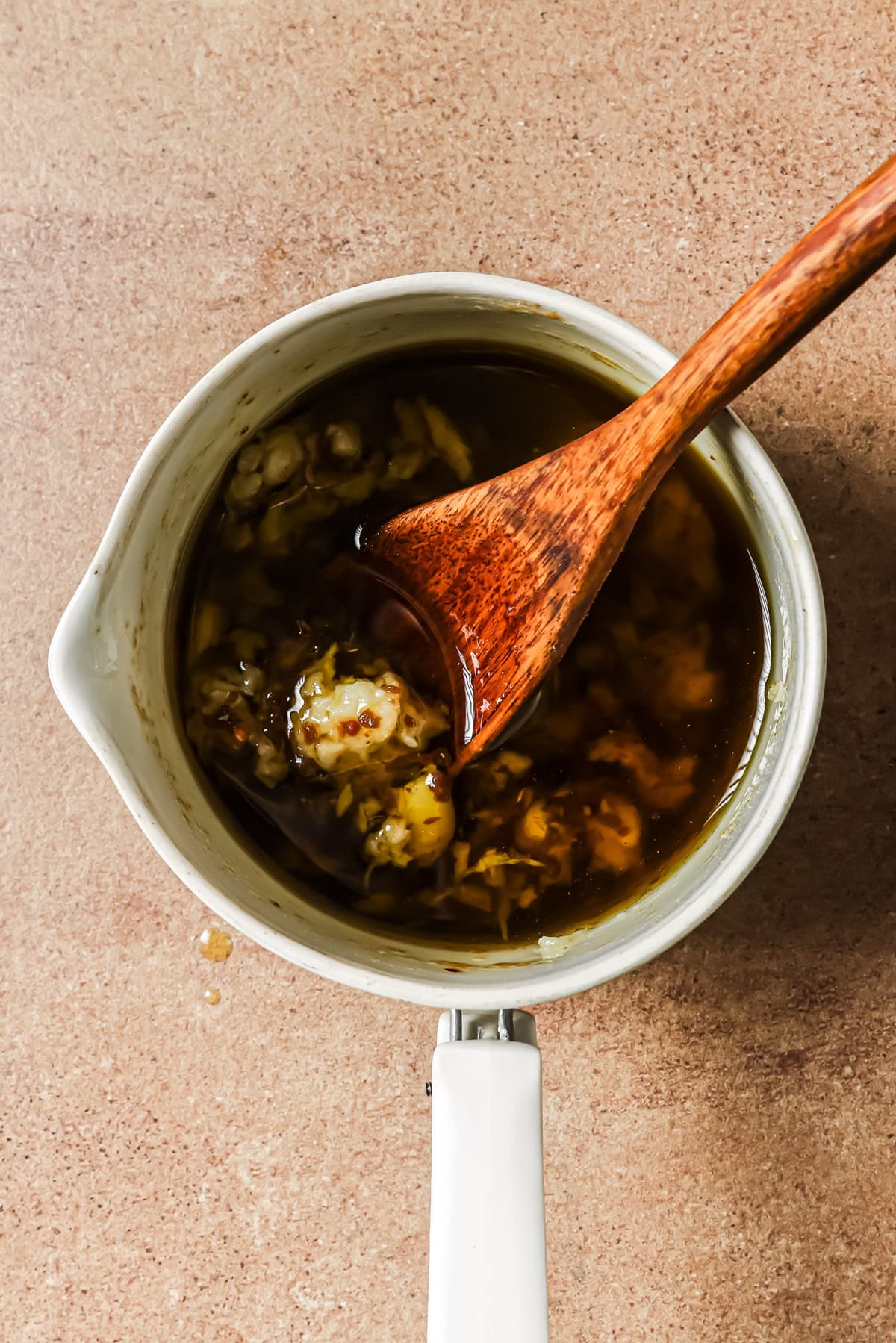 A wooden spoon stirs a dark, garlic-infused sauce in a small white saucepan.