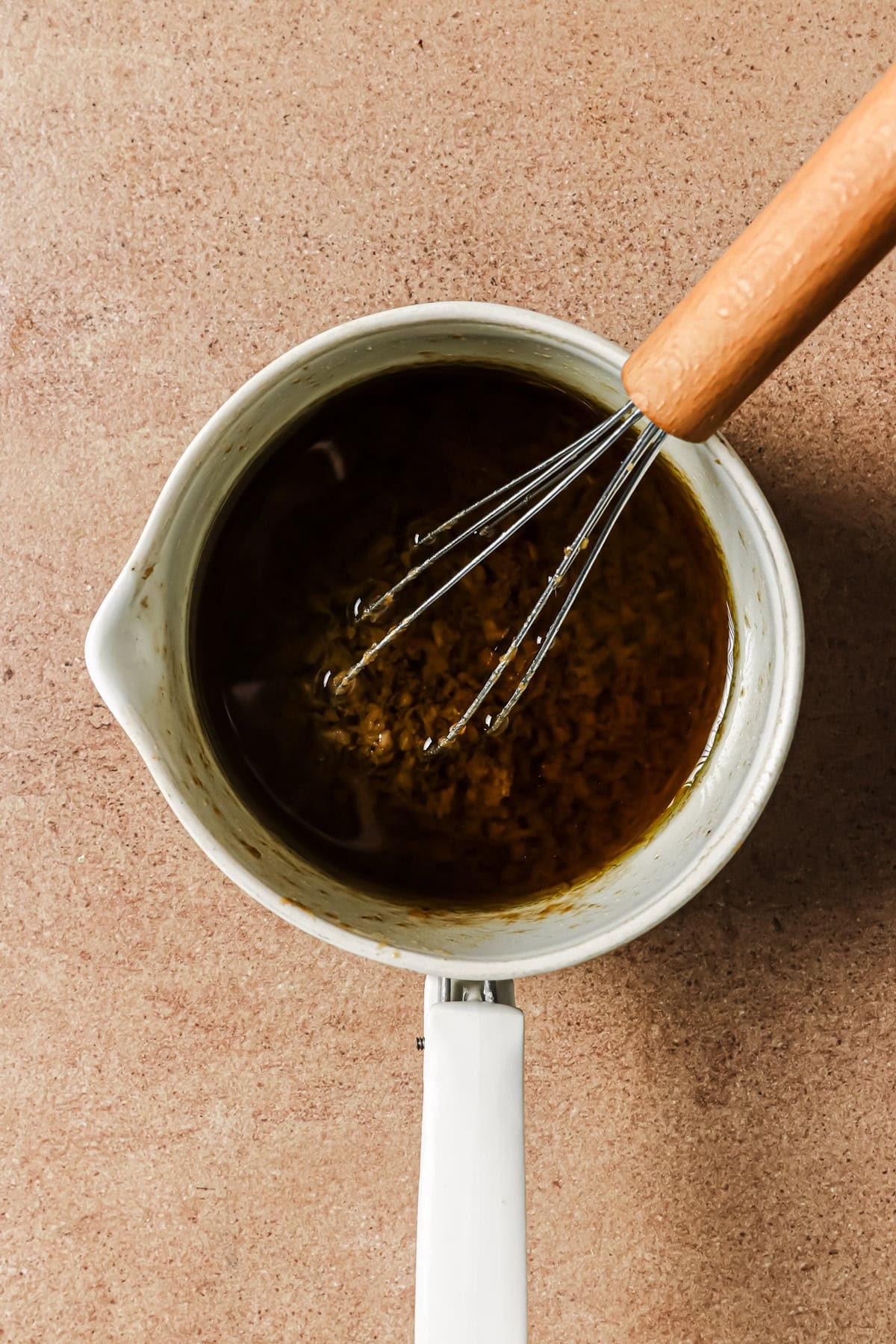 A wire whisk in a saucepan mixing a dark brown liquid with bits of seasoning.
