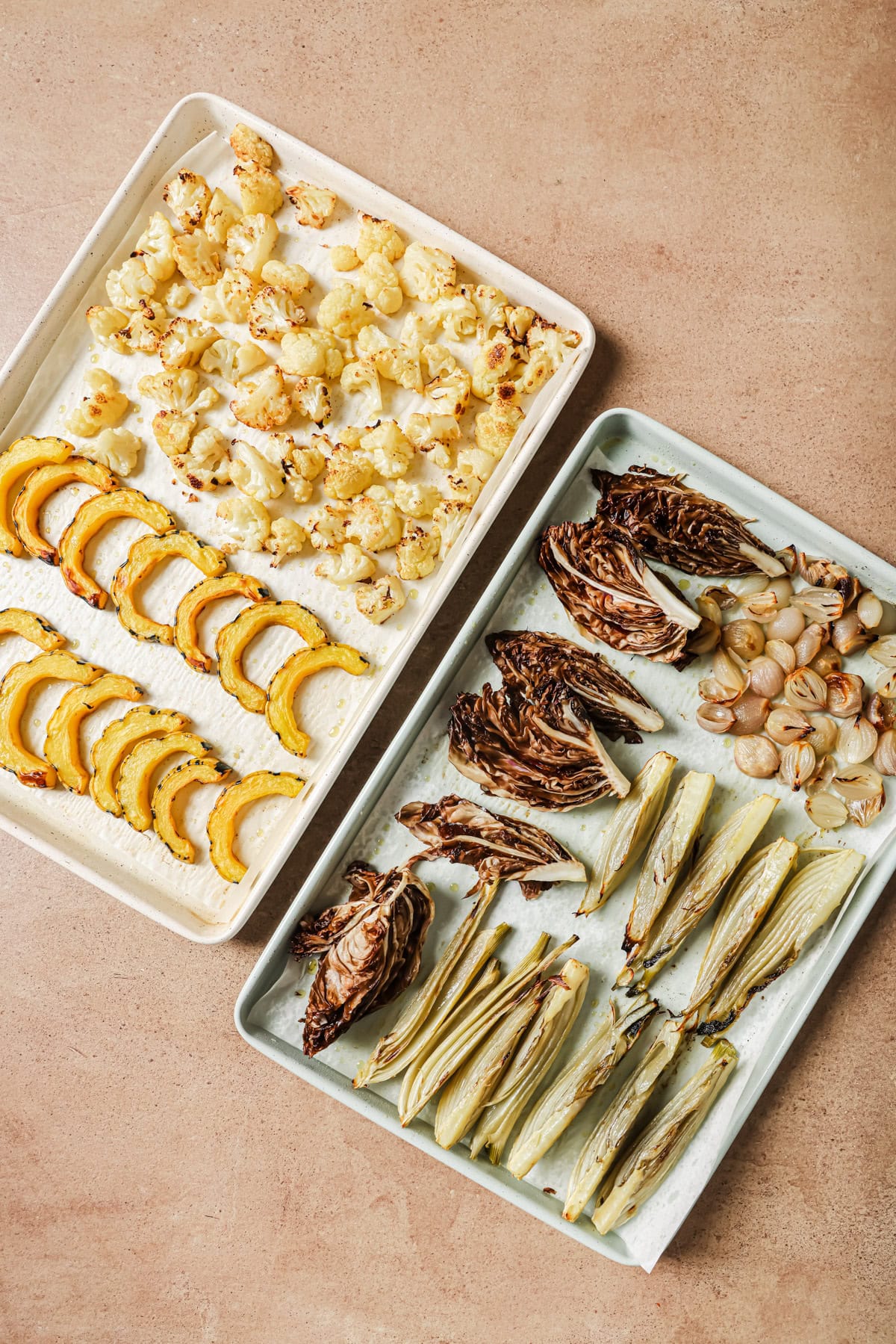 Two baking sheets with roasted vegetables: one with cauliflower florets and squash slices, the other with radicchio wedges, roasted shallots, and fennel stalks.